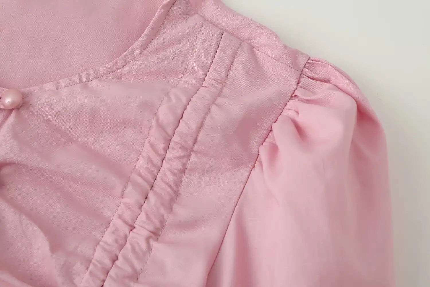 A soft pink women slim-fit long sleeve top featuring a round neckline with gathered fabric and delicate pearl button detail.