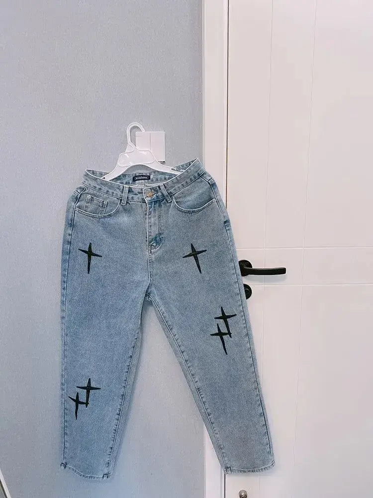 Ce modèle de jeans en denim délavé clair présente une broderie croisée noire sur les jambes et est présenté suspendu à un cintre blanc sur cette image.