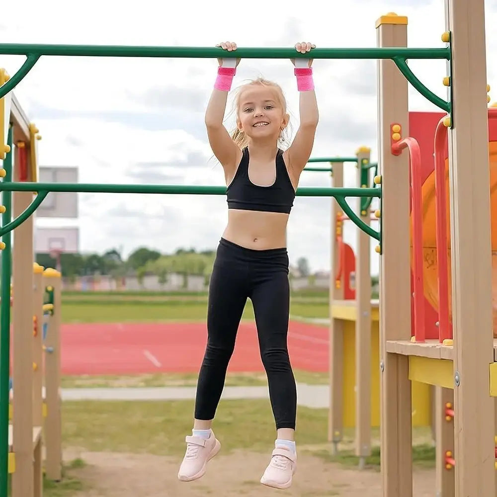 A young girl in black athletic wear hangs from green playground bars wearing pink kids gymnastics wristbands designed for grip and support during exercise.