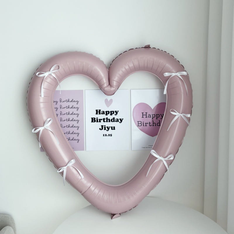 Un marco de foto de globo de lámina con corazón de color rosa suave de 40 pulgadas, adornado con cintas blancas y sujetando tarjetas de cumpleaños.