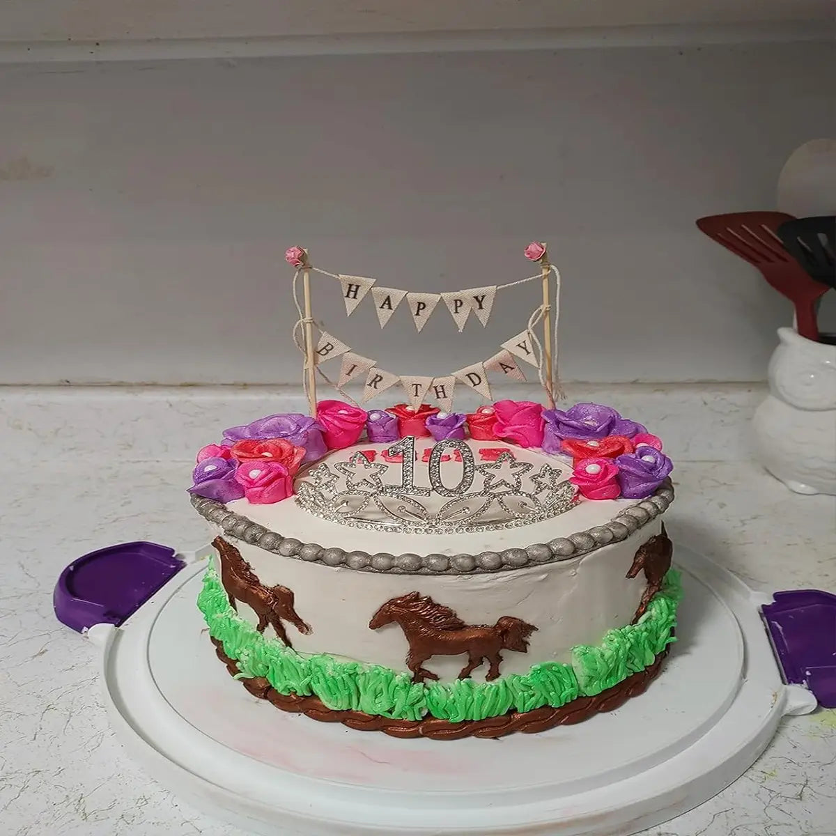 Un gâteau blanc d’anniversaire présentant un fanion de gâteau avec des roses roses, décoré de fleurs en sucre rose et violet, d’un centre de tiara argentée affichant le chiffre 10, d’ornements de chevaux bruns et d’une herbe en glaçage vert.