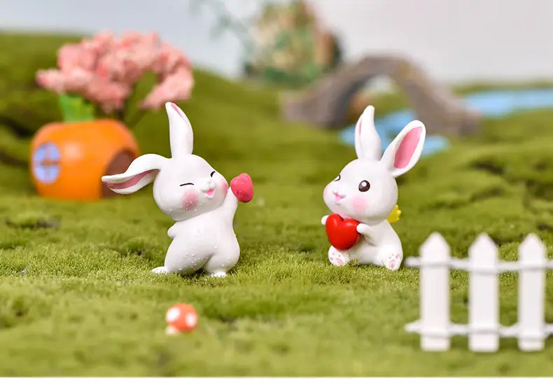Deux petites figurines miniatures en résine céramique blanche de lapin, avec des oreilles internes et des joues roses, l’une tenant un cœur rouge et l’autre offrant un petit objet rose, conçues comme décorations pour jardin féerique.