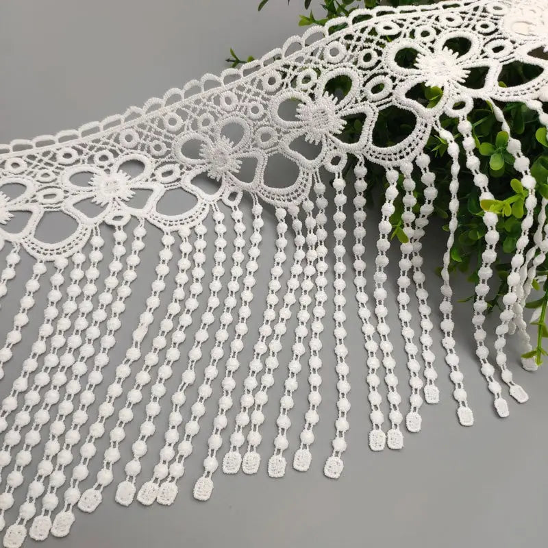 Tissu à bordure de dentelle à perles blanches, présentant une dentelle blanche avec des motifs floraux et un frange de perles.
