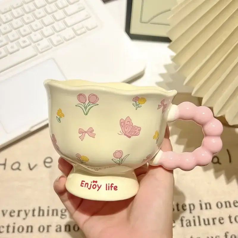 Une tasse tulipe en céramique crème pastel avec une anse rose perlée et de délicates illustrations florales et de papillons, dans une boîte cadeau à motif floral.