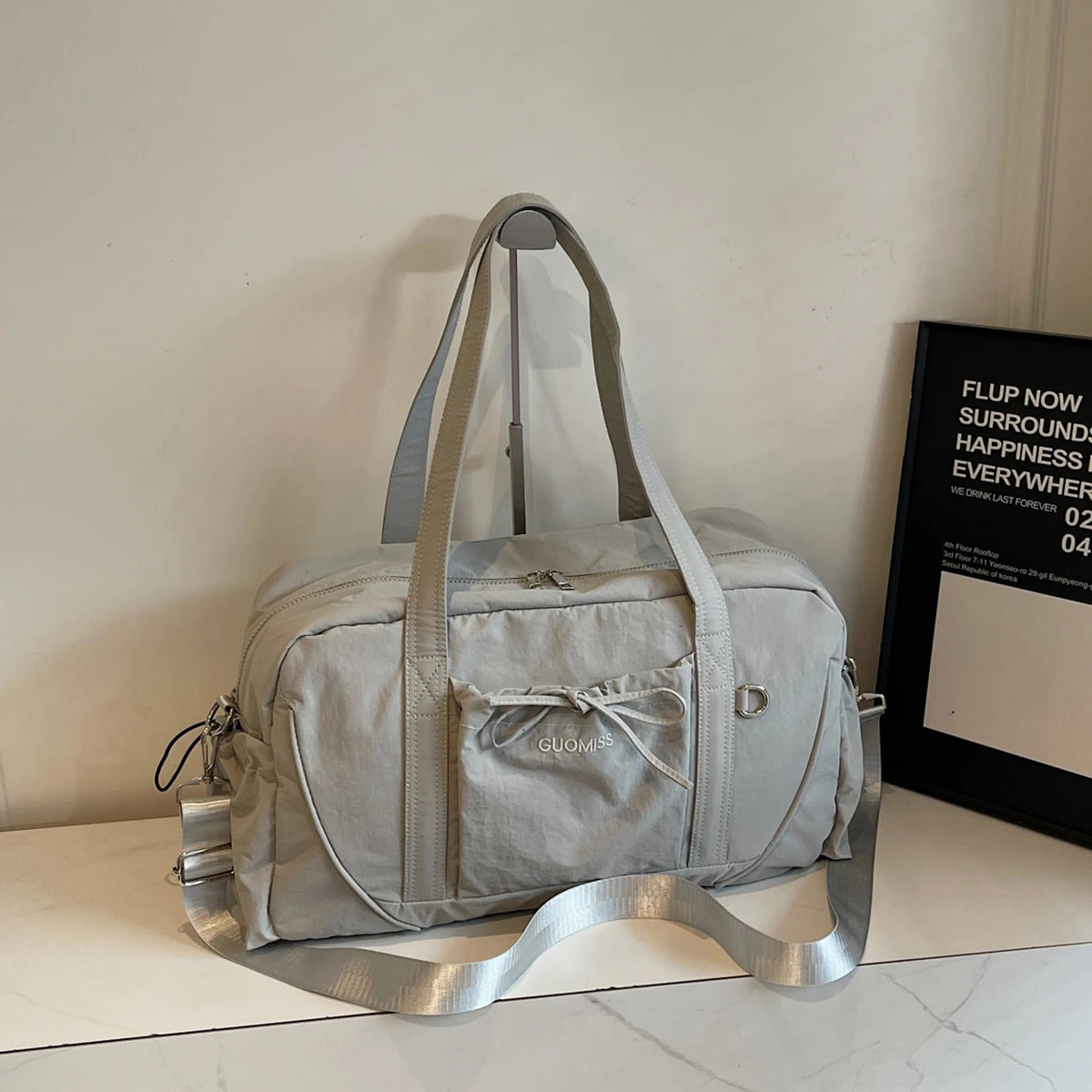 Light gray guomiss large capacity travel duffel bag made of nylon with a silver zipper pull adjustable shoulder strap and a front pocket tied with a bow.