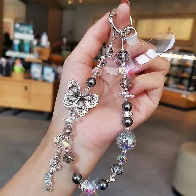 Un porte-clés de chaîne pour téléphone en forme de papillon argenté, orné de perles transparentes et gris foncé, rehaussé d’accents en cristal scintillant, avec une pince en plastique transparent pour un usage avec le téléphone mobile.