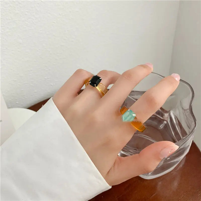A hand wearing two colorful resin crystal geometric ring bands with black and teal gemstones set in translucent amber resin for women.