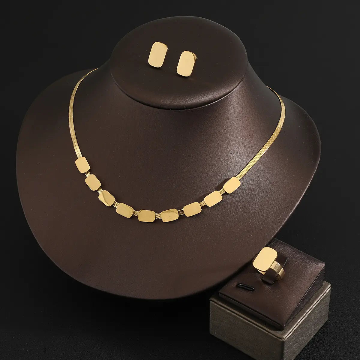 Juego de joyería de acero inoxidable color dorado para mujeres, con un collar con dije rectangular de tono dorado, cadena delicada y aretes a juego, mostrado en un busto de maniquí color marrón oscuro.