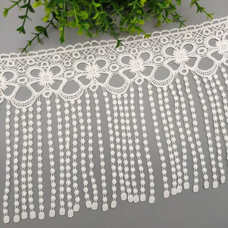 Tissu à bordure de dentelle à perles blanches, présentant une dentelle blanche avec des motifs floraux et un frange de perles pendantes.