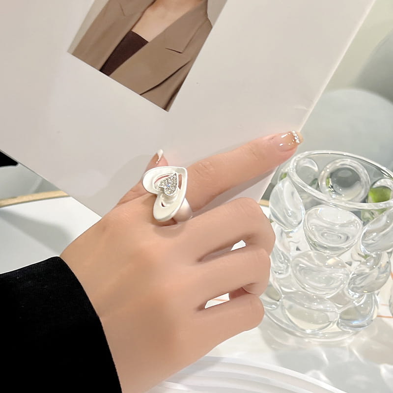 Anillo ajustable con forma de corazón en acabado blanco, con un acabado en tono dorado y un detalle central de pedrería brillante.
