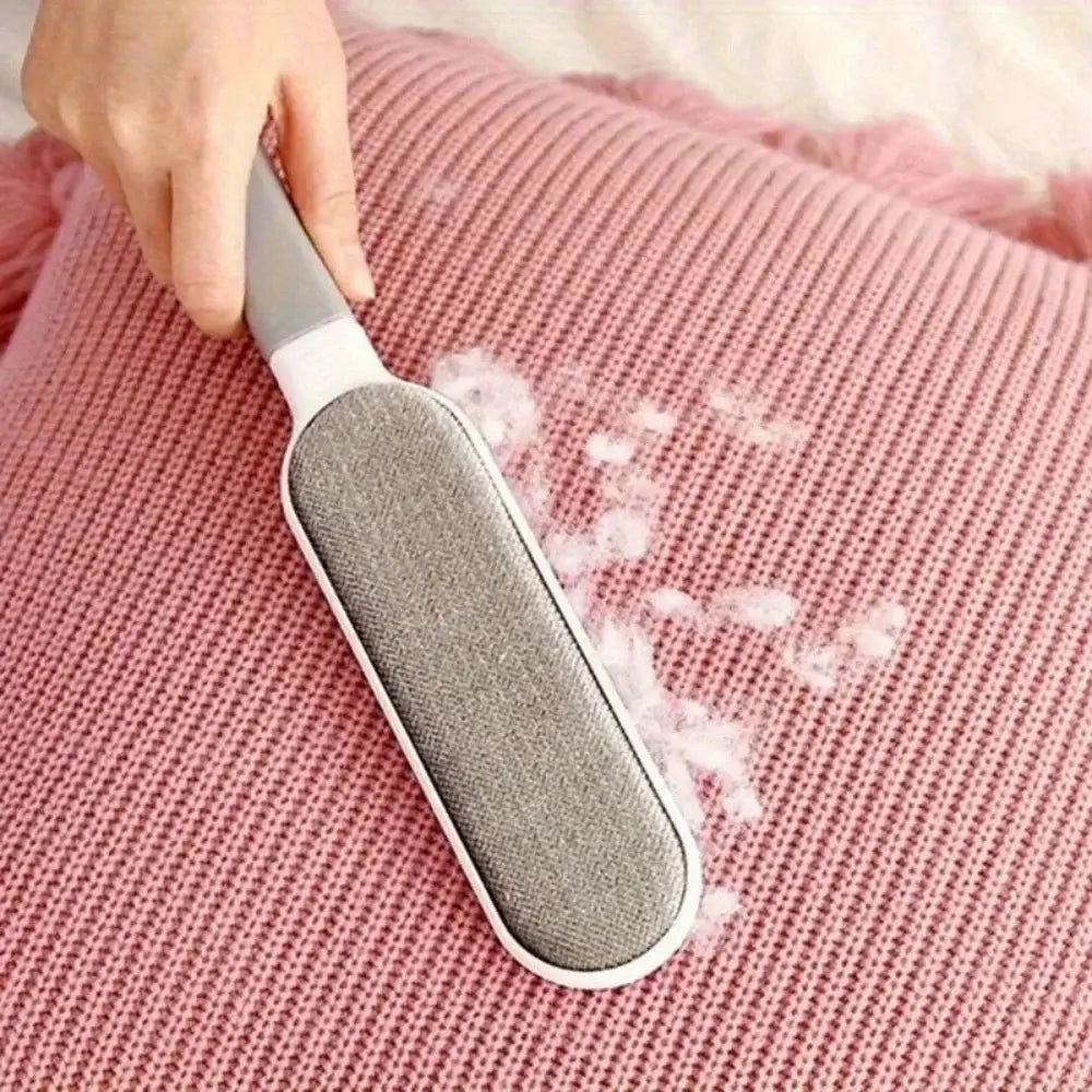 Grauer stoffbezogener doppelseitiger Tierhaar-Entferner und Fusselbürste mit weißem Griff, der verwendet wird, um flauschige Fasern von einer rosa gestrickten Oberfläche zu entfernen.