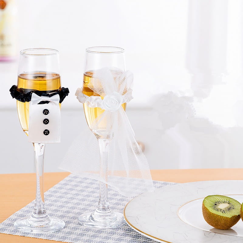 Dos copas de vino de cristal transparentes para la novia y el novio con una cubierta tipo esmoquin en blanco y negro y una delicada cubierta de encaje blanco.