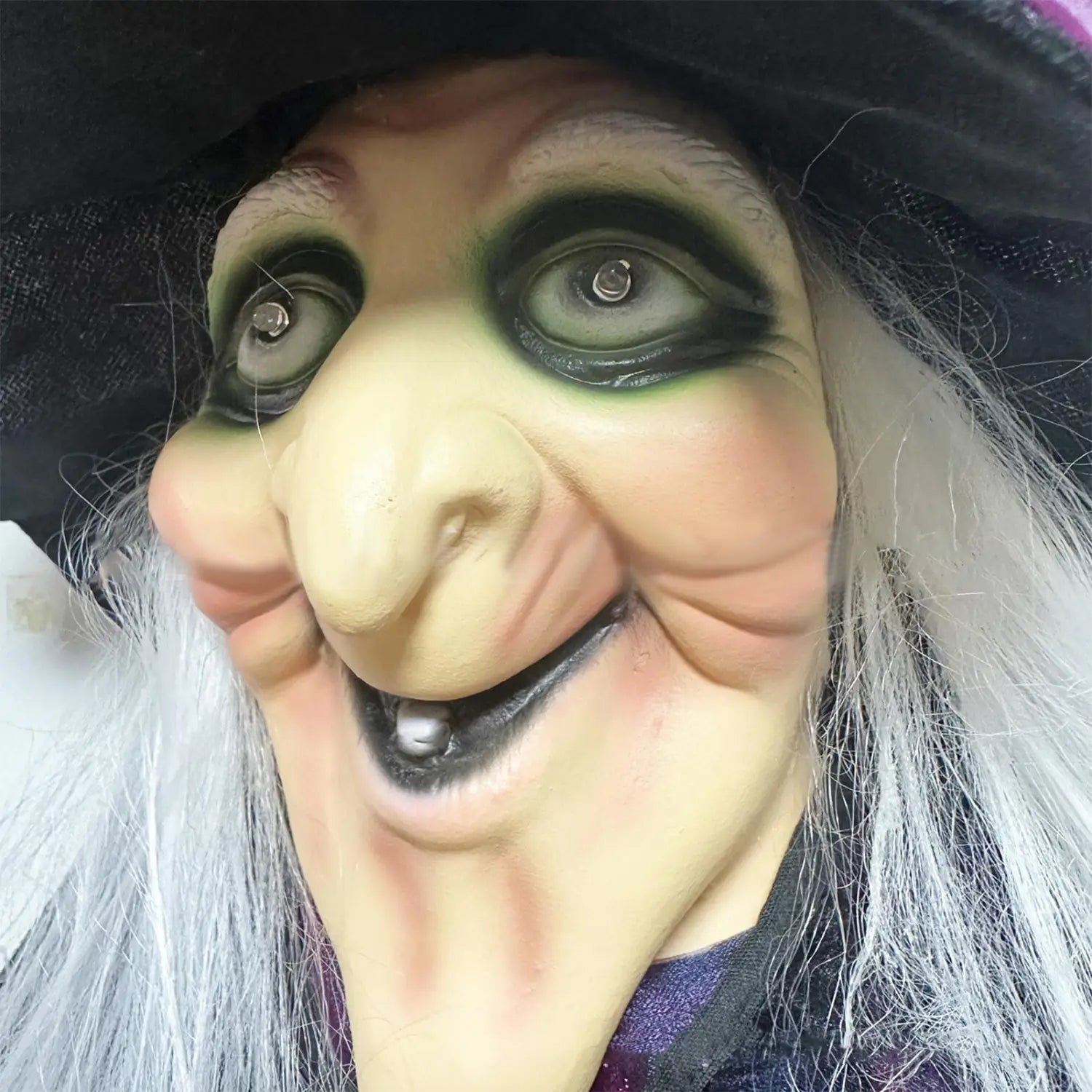 Accessoire de sorcière d’Halloween détaillé et réaliste, présentant un masque à activation sonore avec un teint pâle, un fard à paupières vert foncé, des cheveux synthétiques blancs et un chapeau en tissu noir.