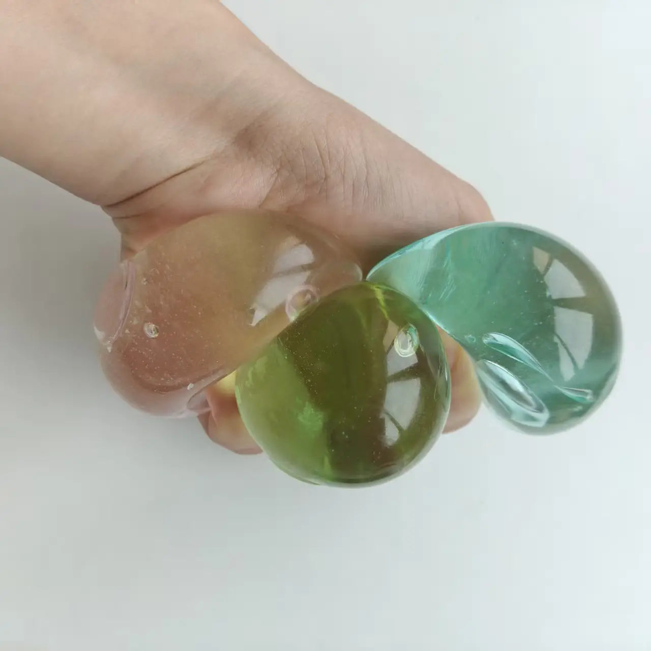 une main tient trois balles anti-stress souples translucides en couleurs pêche pastel, vert et turquoise, montrant la vue de face de la balle anti-stress verte à presser et du jouet sensoriel anti-stress rose vert.