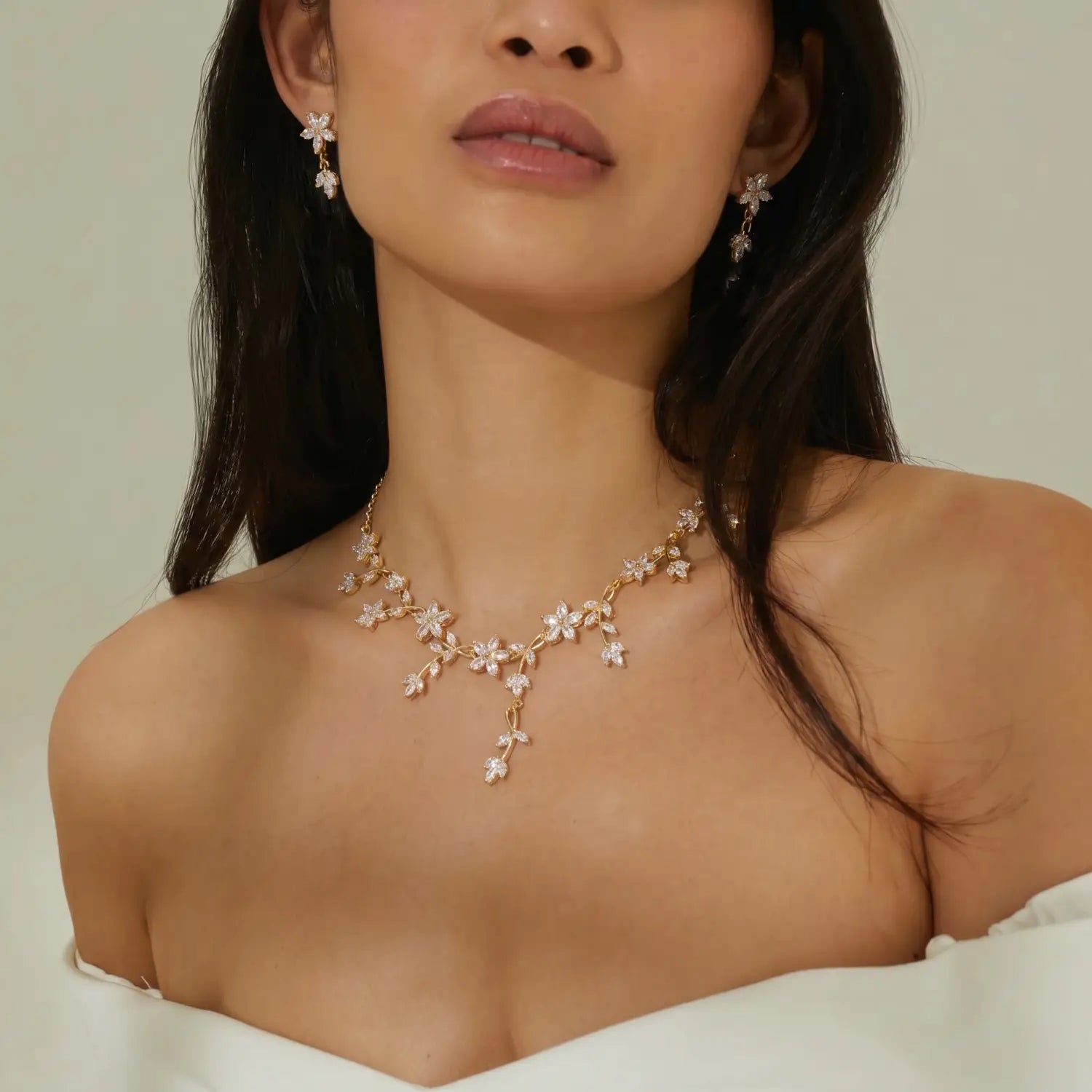 Set aus einer funkelnden Brautkette und Ohrringen mit einer zarten goldfarbenen Kette, geschmückt mit glitzernden, diamantenähnlichen Blumen und Blättern, mit einem zentralen Anhänger, der unterhalb des Schlüsselbeins herabhängt.