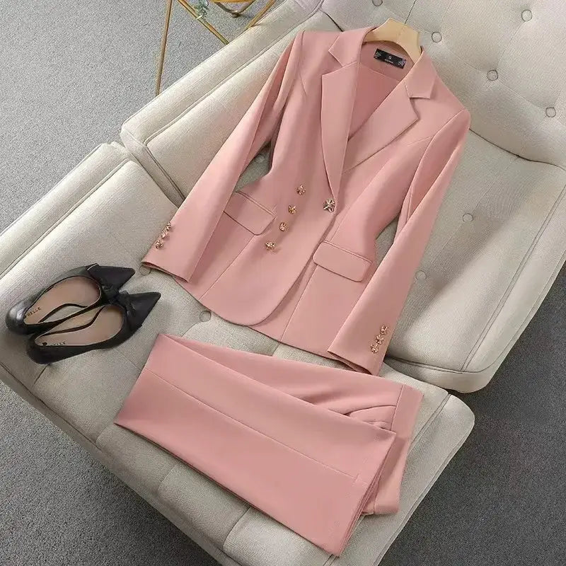 Un ensemble de tailleur d’affaires pour femme cadre sur mesure rose poudré, composé d’une veste avec des accents de boutons floraux à finition ton-or et d’un pantalon assorti, présenté sur un canapé beige capitonné.