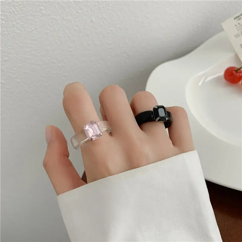 A hand wearing two colorful resin crystal geometric rings for women featuring one translucent band with a pink rectangular gemstone and one black band with a matching black rectangular gemstone.