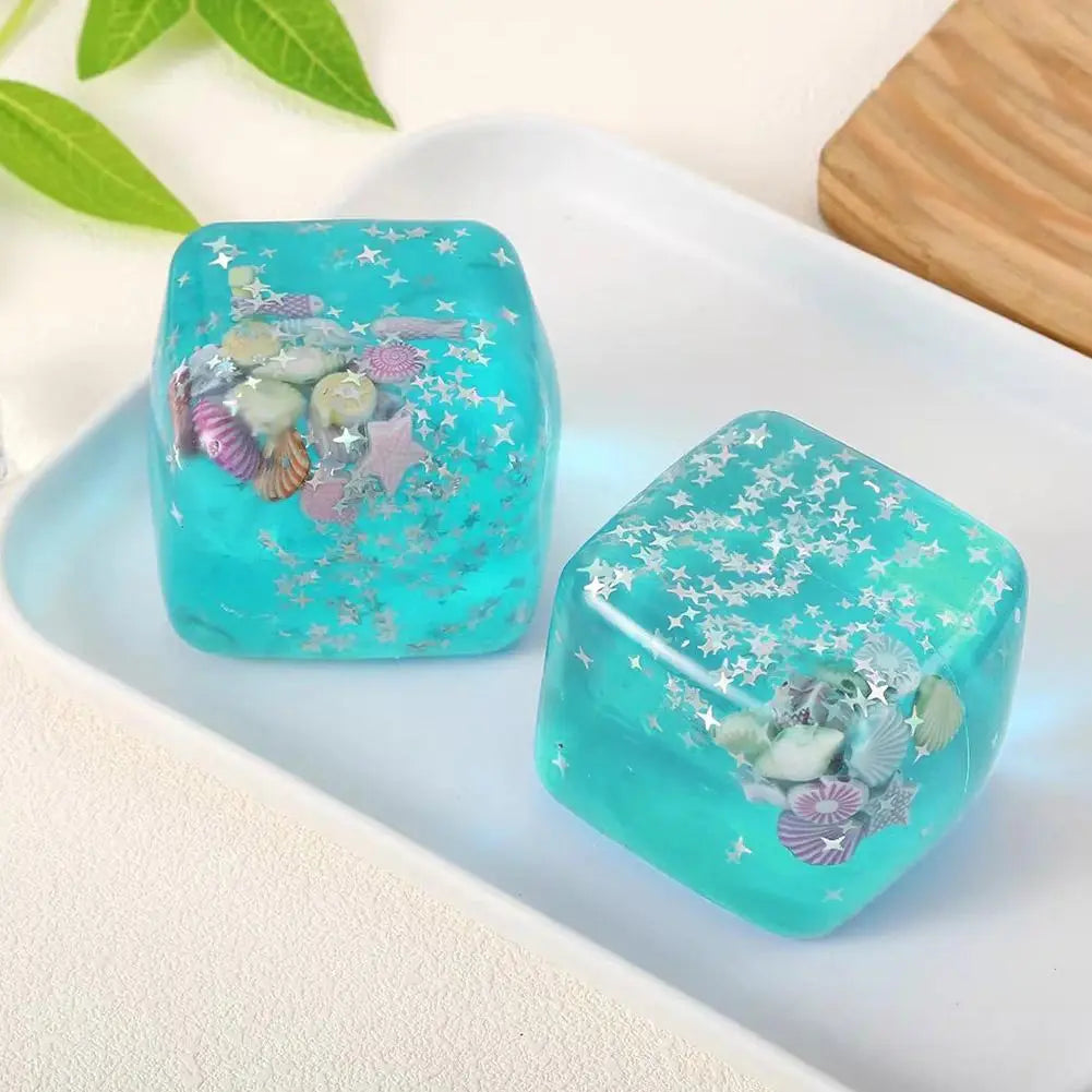 Zwei durchscheinende türkisfarbene Harz-Fidget-Würfel, mit eingearbeiteten Miniaturmuscheln und silbernem sternförmigem Konfetti, von vorn als nachdenkliches Geschenk dargestellt.