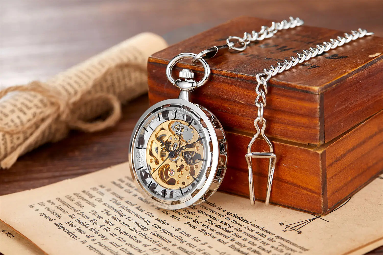 Montre de poche mécanique à tonalité argent avec mécanisme d’engrenages squelette doré visible et indications en chiffres romains, posée sur un livre ouvert à côté de sa chaîne assortie.