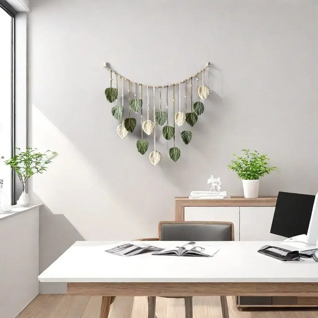 A delicate macrame leaf wall hanging boho decor featuring a curved arrangement of green and cream-colored leaf-shaped ornaments suspended from thin cords.