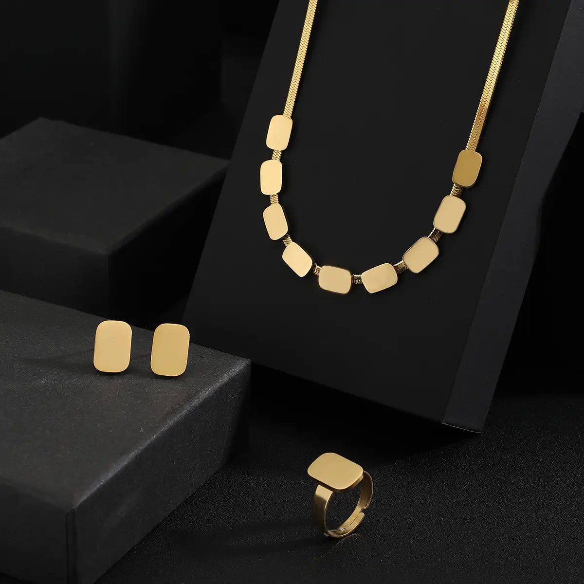juego de joyería de acero inoxidable color dorado para mujeres, con un collar de dije rectangular de tono dorado, aretes de cadena a juego y anillo.