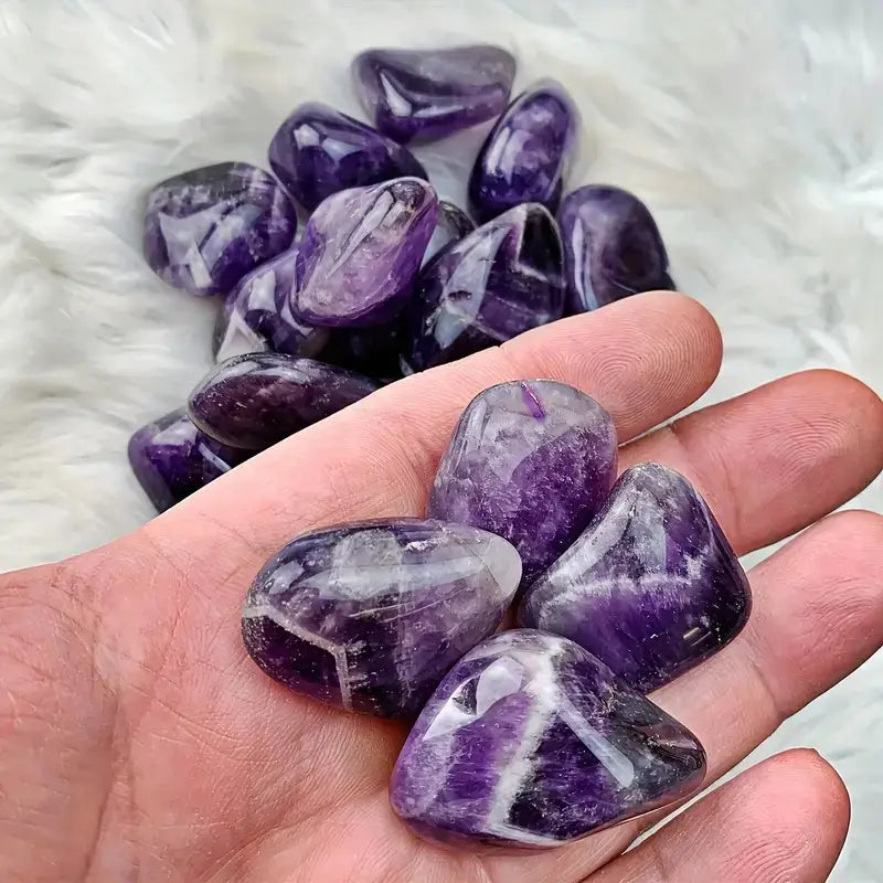 Une main tient plusieurs pierres roulées naturelles d’améthyste polies et lisses, pesant 100 g, aux teintes pourpre profond et veines blanches, utilisées pour des soins de reiki.