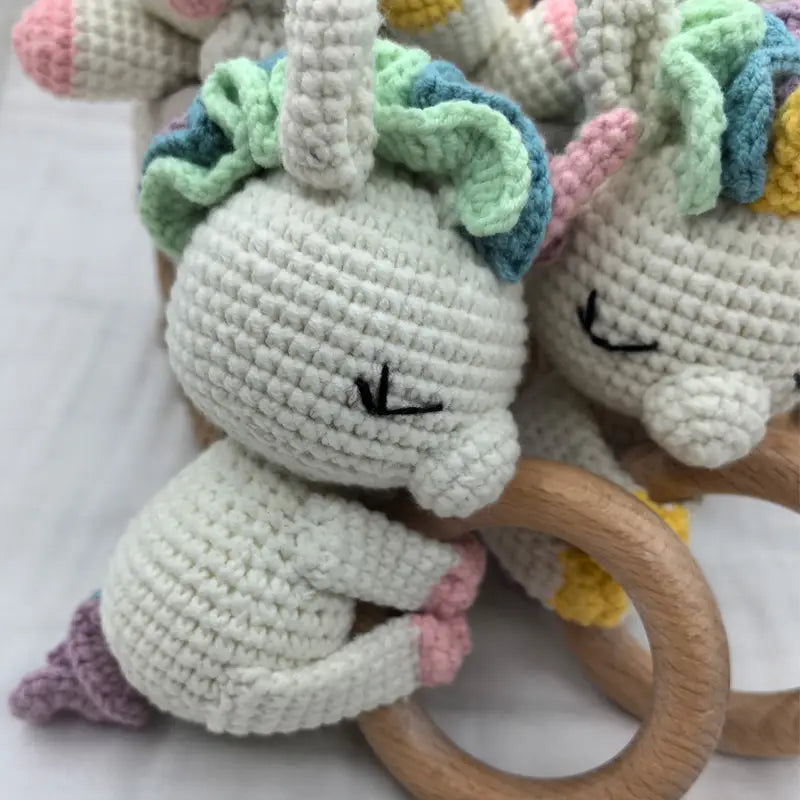 Niedliche weiße gehäkelte Einhorn-Baby-Schmusedecke-Sicherheitsdecke-Rassel mit pastellgrüner und blauem Mähnenhaar, pinkem Horn und Schwanz, die einen hölzernen Ring hält.