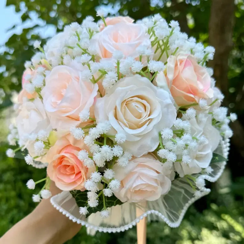 A delicate white silk rose wedding bouquet featuring peach and cream artificial roses intertwined with white babys breath and wrapped in a sheer lace ribbon with pearls.