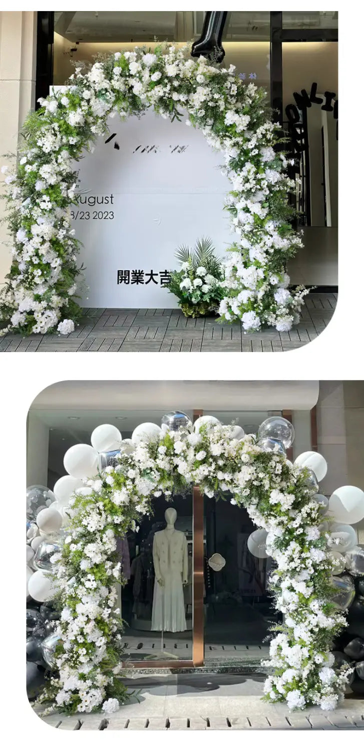 Un exuberante arco floral blanco con soporte adornado con rosas blancas artificiales, vegetación verde y globos transparentes que enmarcan una entrada de local.