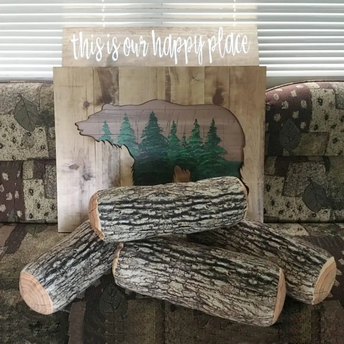 rustic wooden log throw pillow with realistic wood grain texture featuring a painted bear silhouette filled with green pine trees and a smaller plank reading this is our happy place in white script