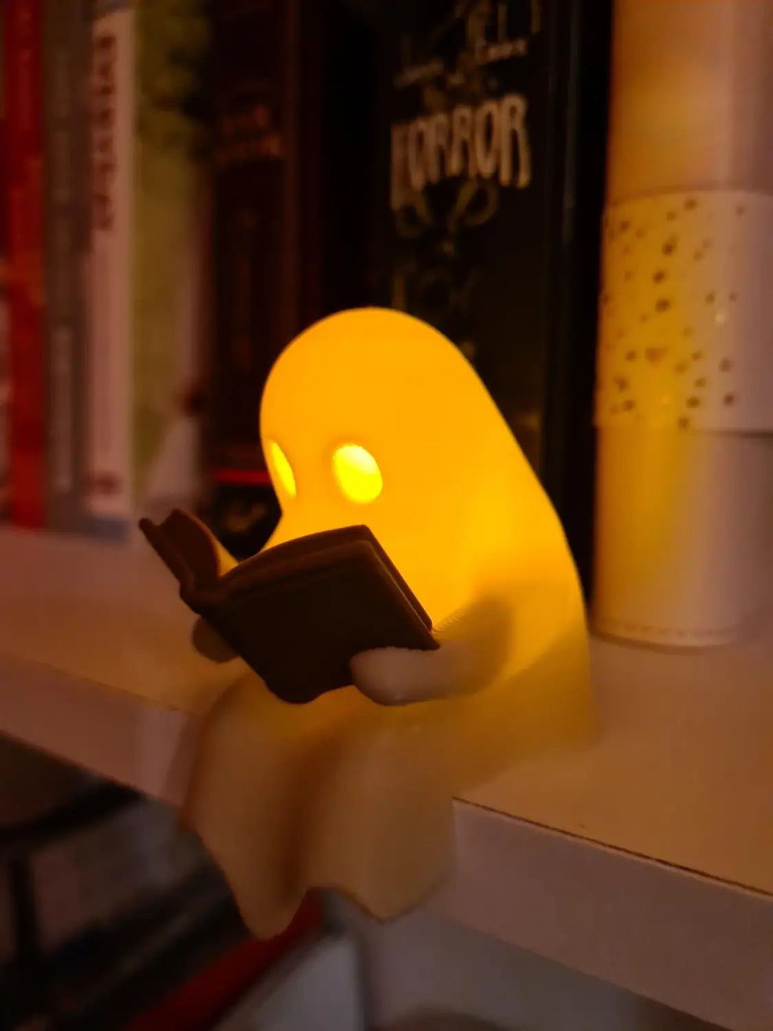 Eine leuchtend gelbe, geisterförmige Lese-Buchfigur als Lampenfigur mit leuchtenden Augen, die ein kleines Buch hält, mit einem flackernden Teelicht.