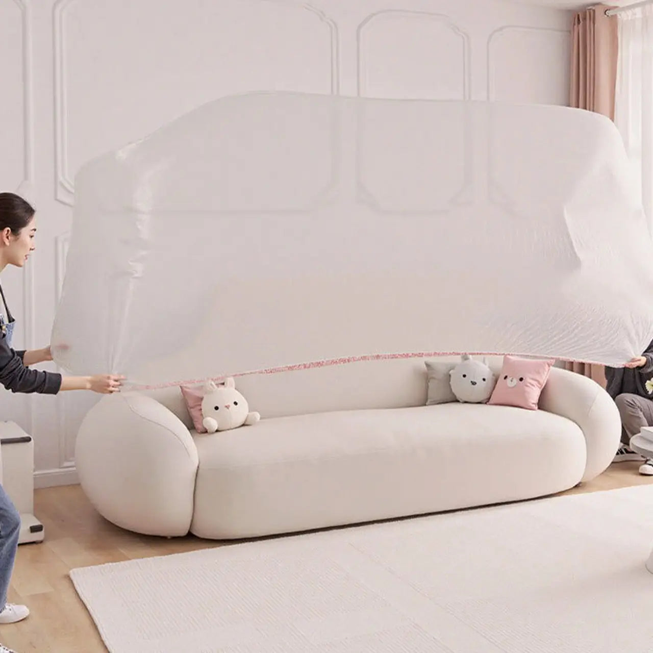 Housse de protection transparente en plastique pour meubles protégeant un canapé en peluche de couleur crème, avec des accoudoirs arrondis et des coussins doux, agrémentés de trois adorables coussins en peluche.