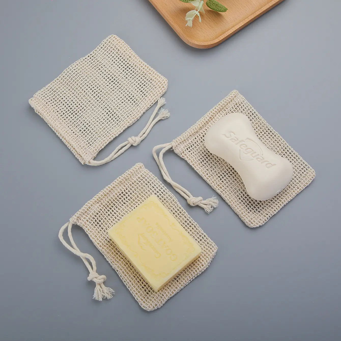 three beige cotton soap saver bags with drawstrings each containing a white embossed and a pale yellow bar of soap in a cotton soap saver bag 3 pack