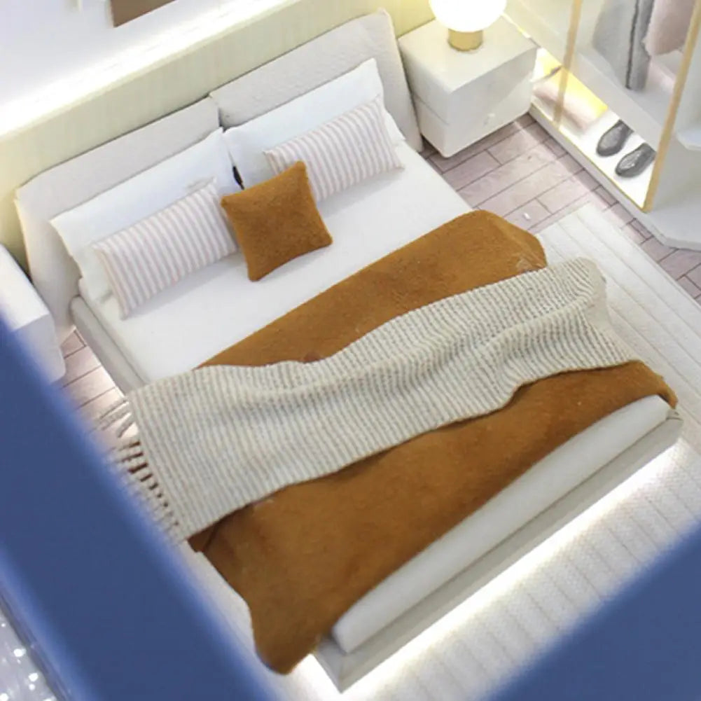 Miniature dollhouse double bed with white bedding cream style featuring white pillows and a brown throw blanket draped over it.