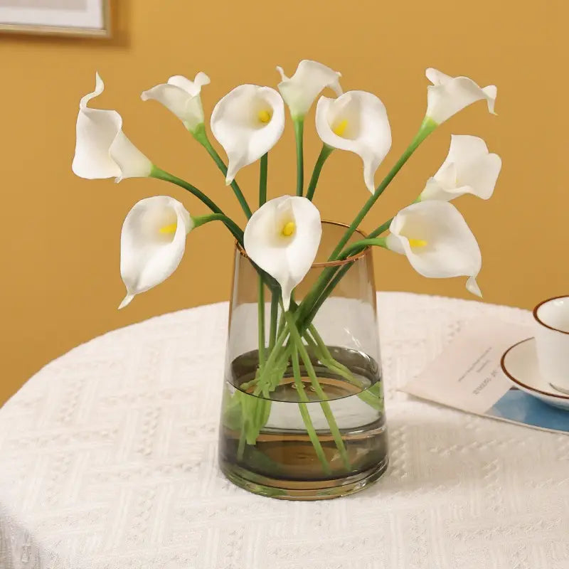 A clear glass vase with a gold rim holding a bouquet of white calla lilies for home decor.