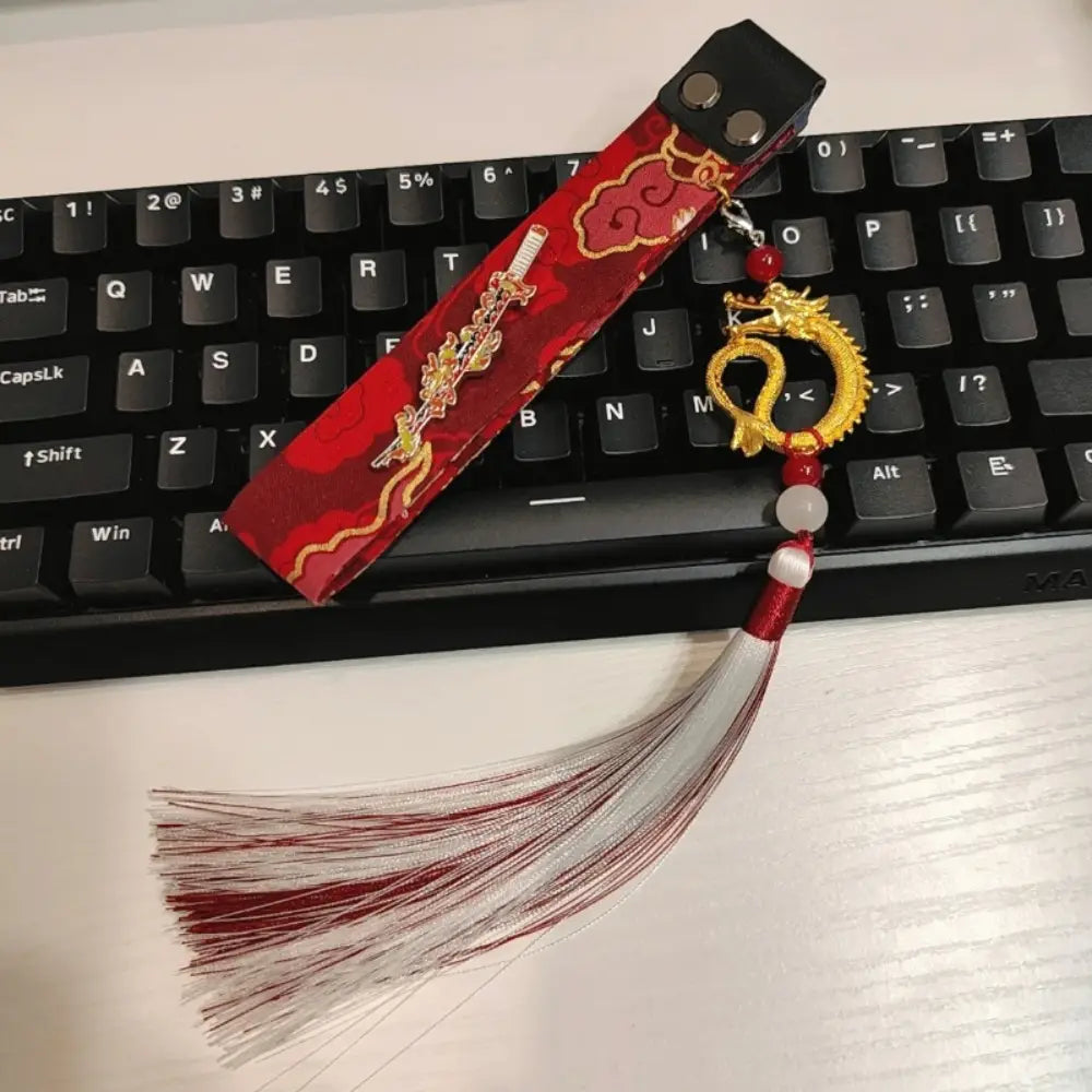 Fourreau d’épée en tissu rouge avec broderie dorée et pompon assorti à breloque de dragon, présenté sur la sangle personnalisée du modèle Ribbon Dragon du clavier Wooting.