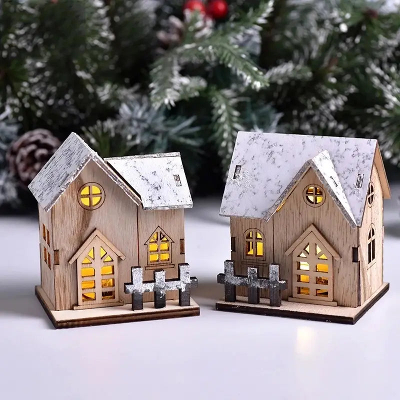 Zwei Miniatur-Holz-Weihnachtshäuser mit schneebestäubten Dächern und leuchtend gelben Fenstern, von denen jedes einen kleinen dekorativen Zaun aufweist; es handelt sich um leuchtende Weihnachtsdekorationen mit LED-Beleuchtung.