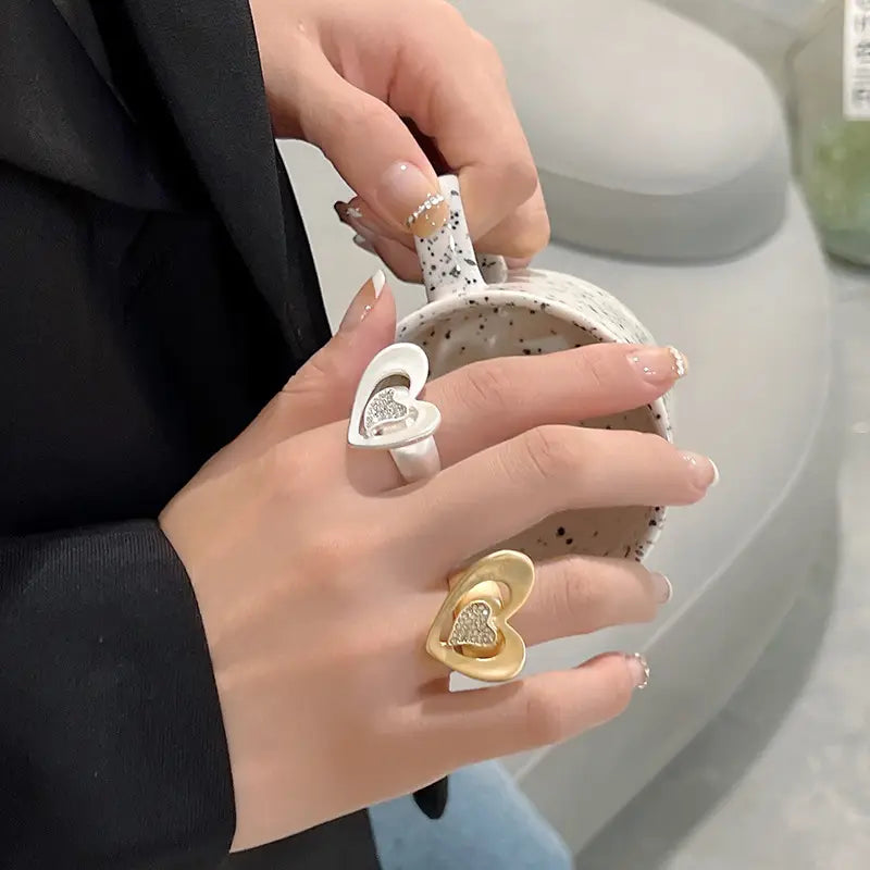 Dos anillos con forma de corazón: uno con aro en tono blanco con un centro de pedrería brillante y otro anillo ajustable en tono dorado con diseños en capas.