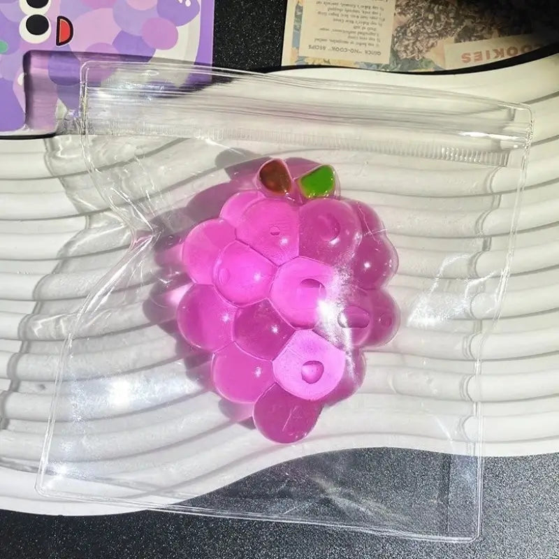 Ein transparenter, rosafarbener Trauben-geformter Taba-licious Fun Fruit-Stressball mit einem grünen und braunen Stiel aus weichem, knetbarem Material, in einer transparenten Kunststoffverpackung, entworfen als lustiges Frucht-Taba-Fidget-Spielzeug