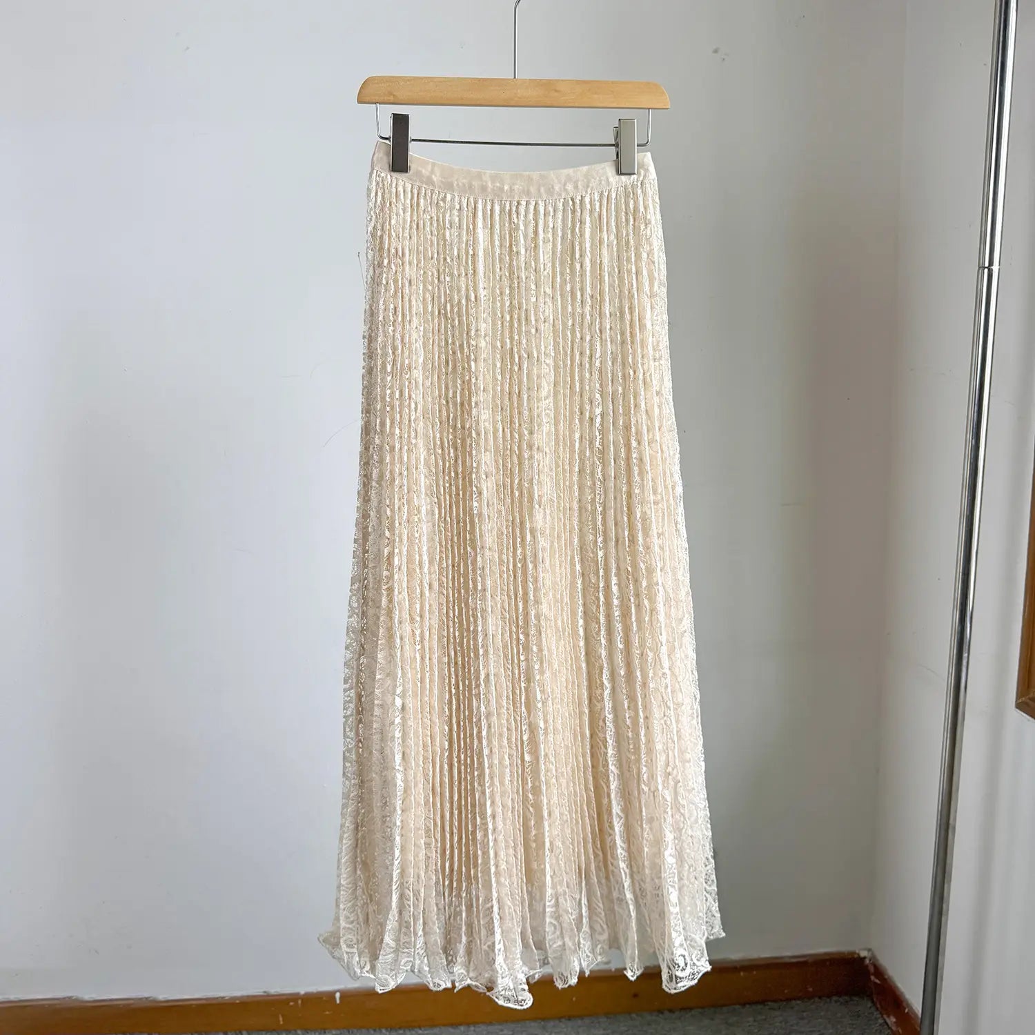 A delicate cream-colored high waist pleated midi skirt made of lace hanging on a wooden hanger for Y2k streetwear.