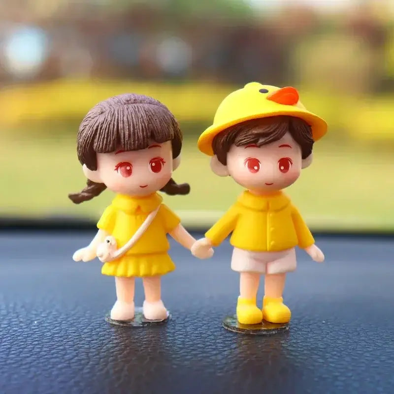 黄色いレインコートを着たカートゥーンのフィギュアが手をつないでいる様子をあしらった、かわいいカップルのカー・ダッシュボード用レジンオーナメント。片方はアヒルの帽子をかぶり、もう片方は小さな白い袋を持っている。