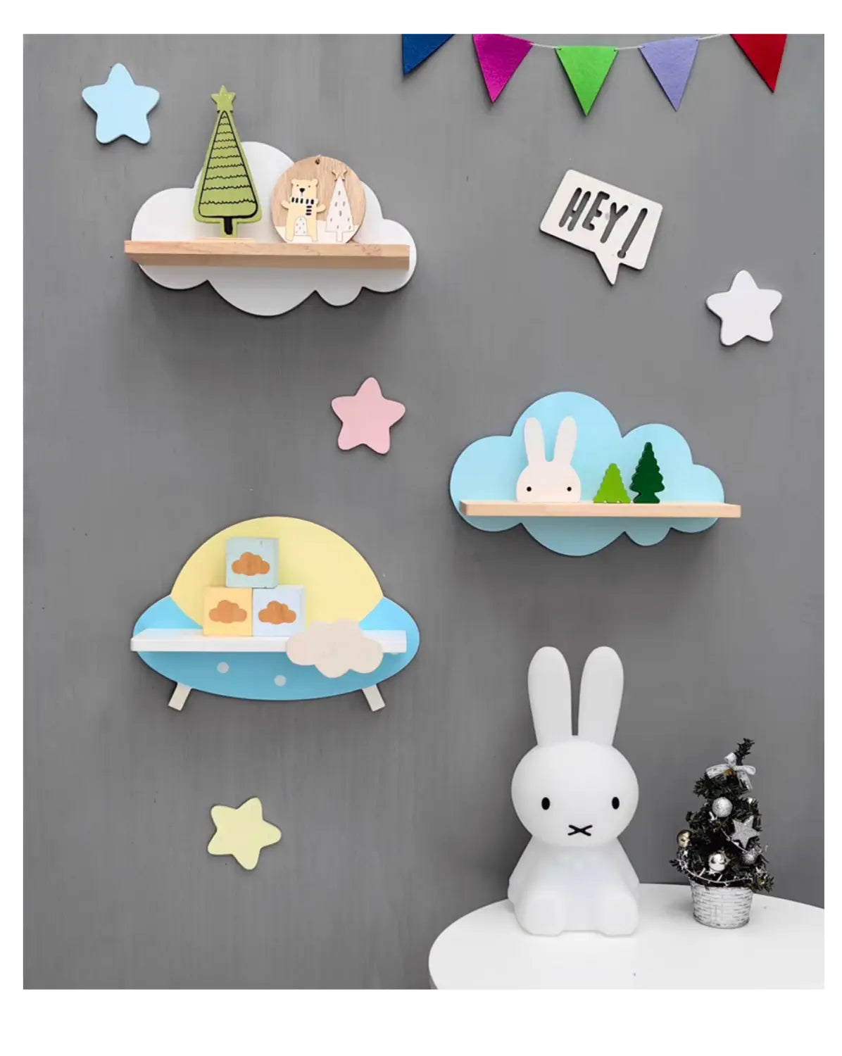 Eine weiße Miffy-Figur mit langen Ohren und einfachem schwarzem Gesichtsausdruck, die auf einer weißen Oberfläche sitzt, neben einem Miniatur-Weihnachtsbaum neben einem Regenbogen-Wolken-Bücherregal aus Holz für Kinder an der Wand.