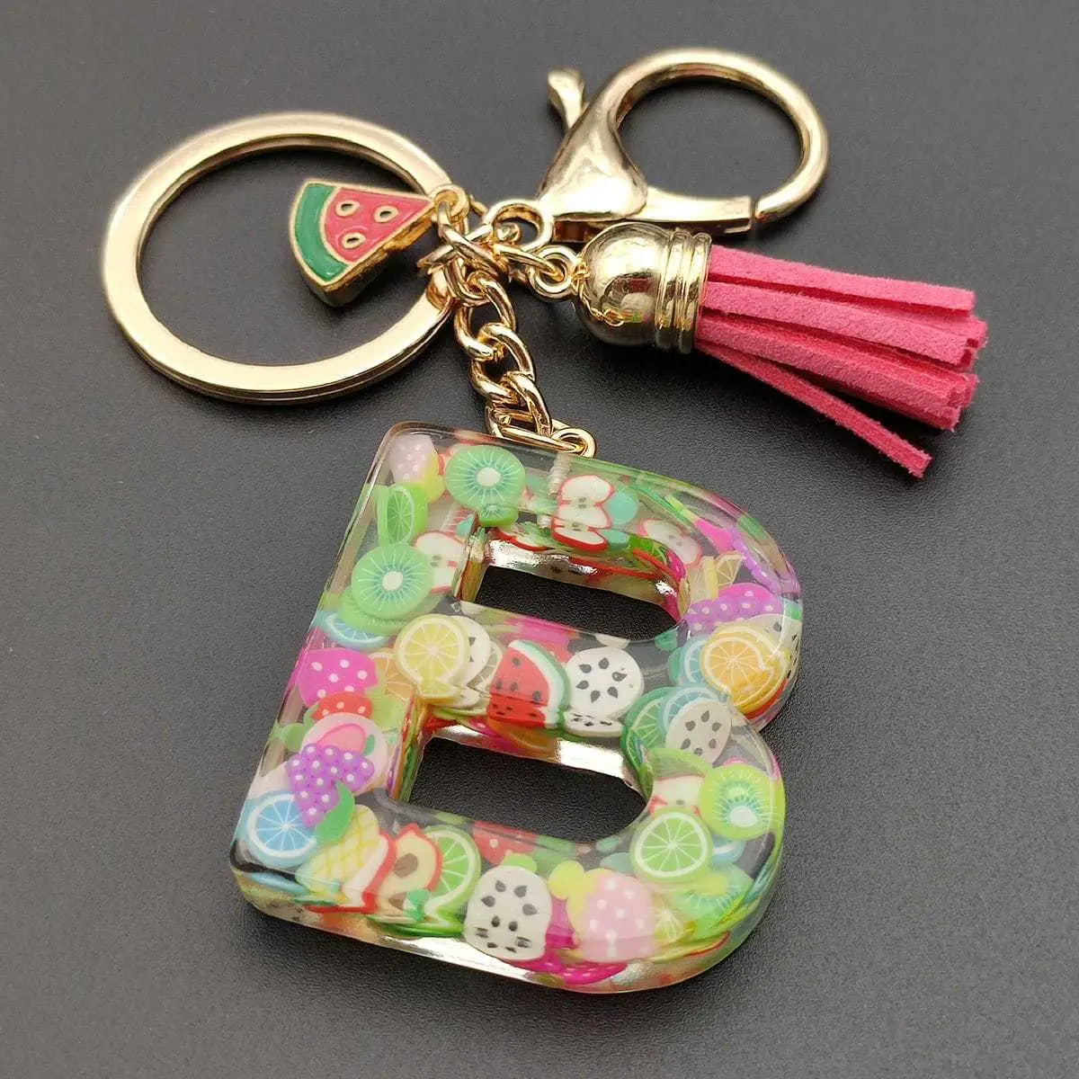 A vibrant transparent resin keychain shaped like the letter b with colorful miniature fruit slices embedded and featuring a gold-toned clasp with a pink tassel.