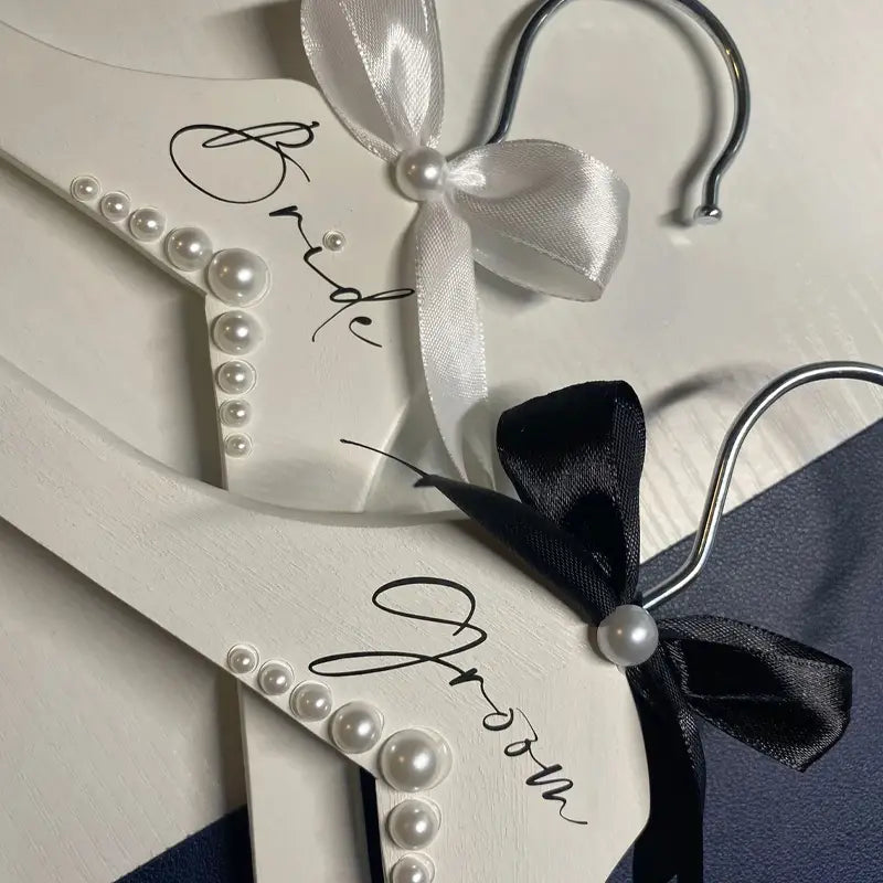 Ensemble de cintres de mariage en bois blanc pour la mariée et le marié, en perles, comprenant des cintres rehaussés de perles avec des nœuds en satin et un cintre étiqueté « mariée » en écriture cursive noire.