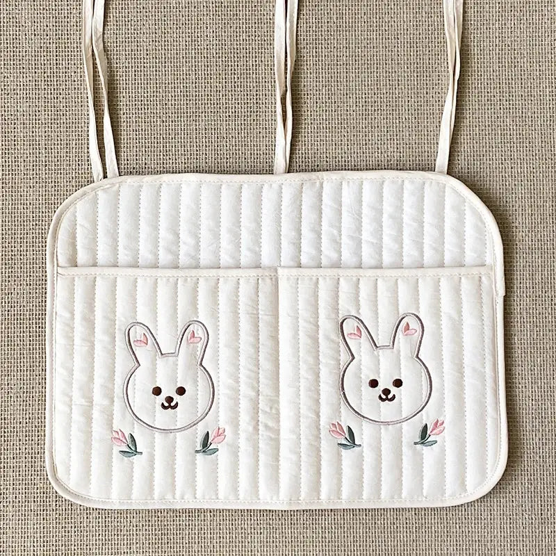 クマの刺繍が施された白いキルティング製ベビーカー用オーガナイザー袋で、刺繍のうさぎの顔とピンクの花が飾られたキルティングのカーシートクッションが特徴です。