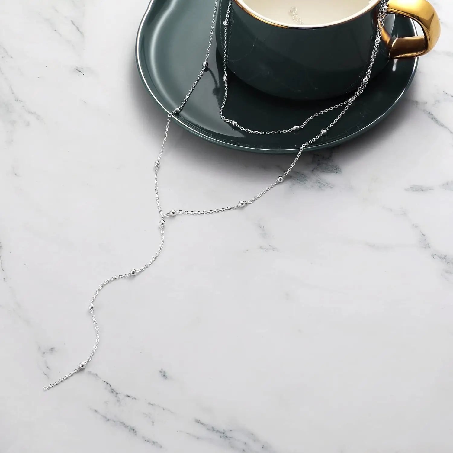 Collier en « y » à superpositions pour femmes, présentant un collier à chaîne superposée à perles en argent reposant sur un petit support en forme de soucoupe vert foncé, à côté d’une tasse assortie.