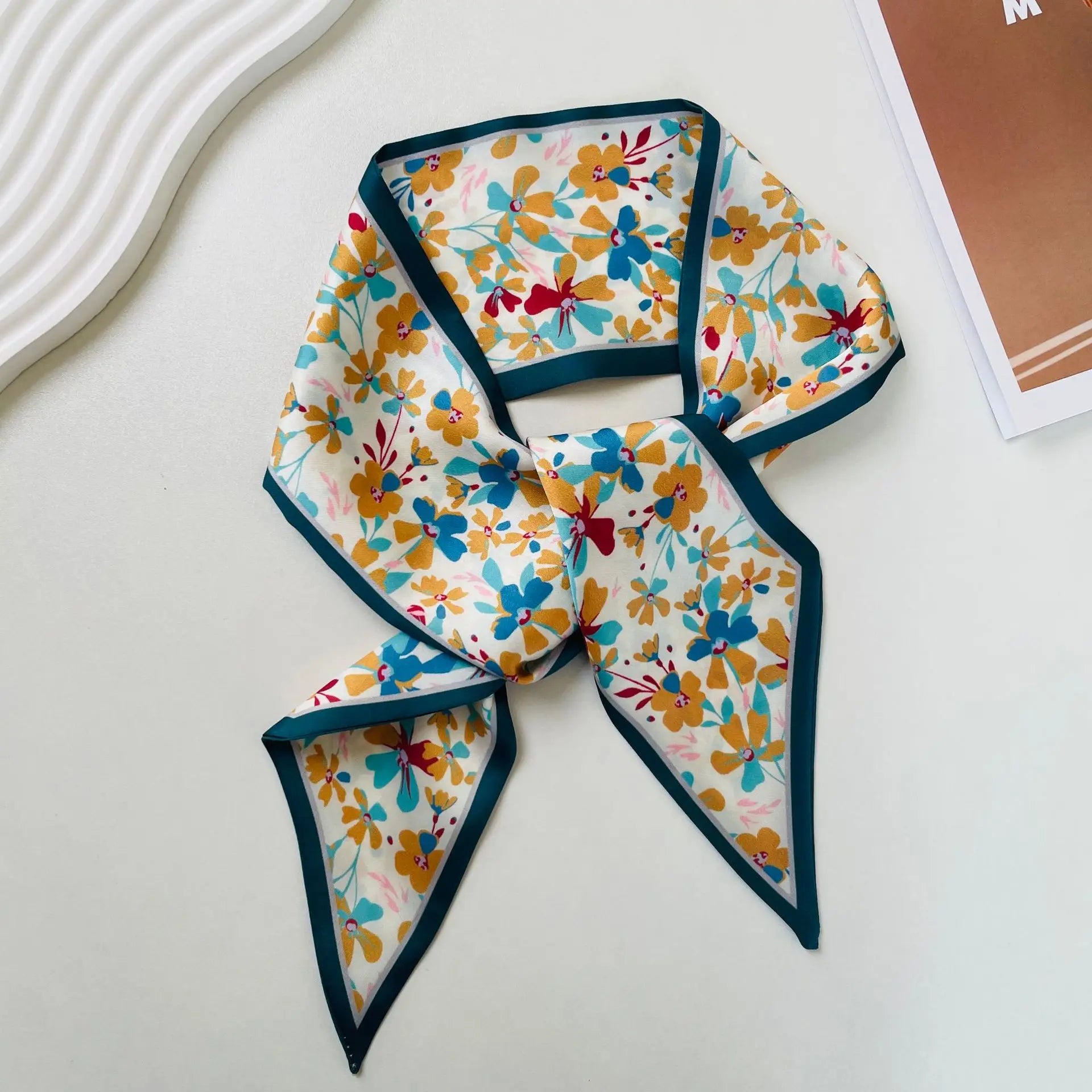 Pañuelo de seda con estampado geométrico, con escote cuadrado, que presenta un patrón floral vibrante con flores en amarillo, azul y rojo sobre un fondo blanco y un borde turquesa elaborado en seda.
