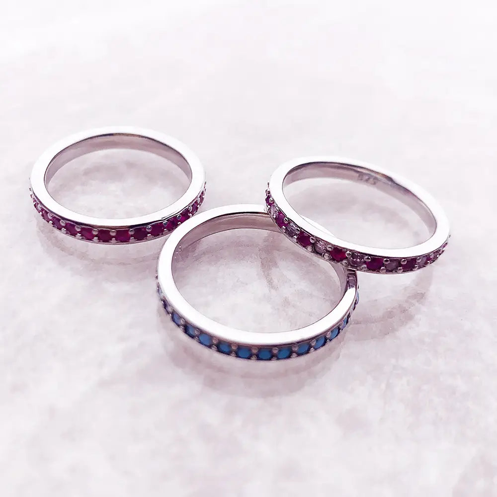 Trois élégantes bagues empilées en argent sterling, chacune ornée d’une ligne continue de pierres précieuses rouges et bleues vibrantes.