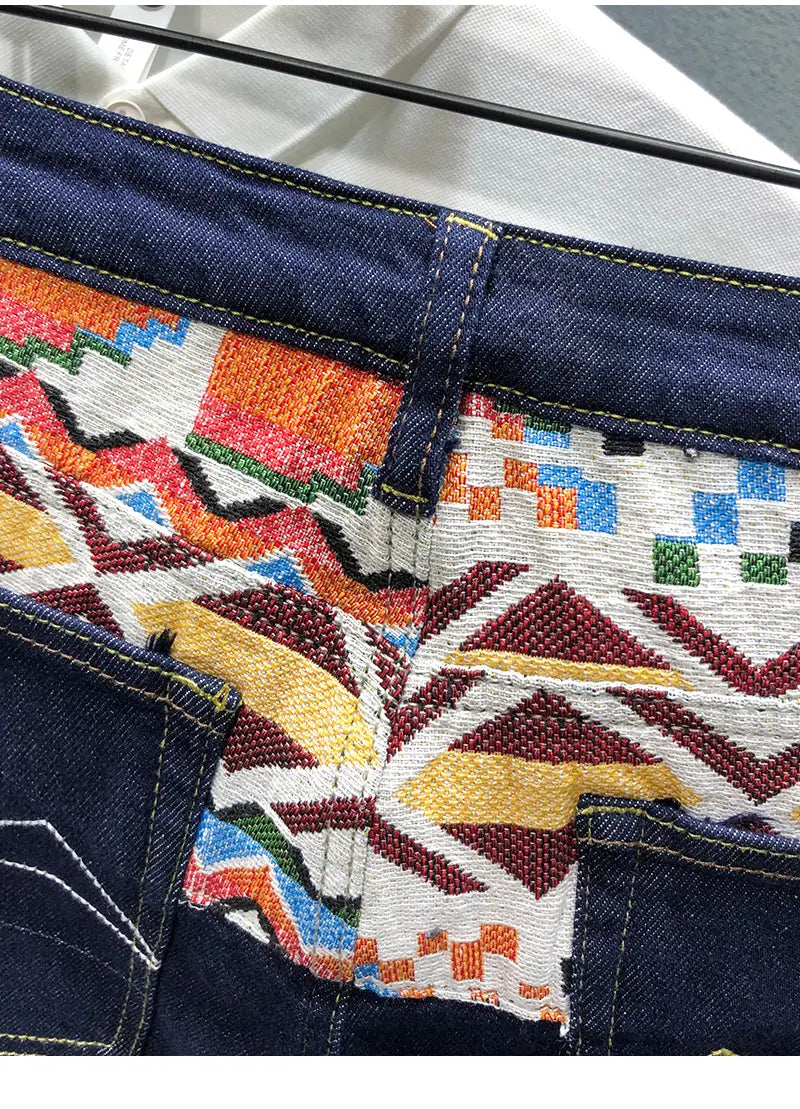 Men slim fit stretch white denim jeans with dark blue sections featuring vibrant multicolored geometric patchwork embroidery including bold zigzags and checkered patterns in orange, red, yellow, green, and white.