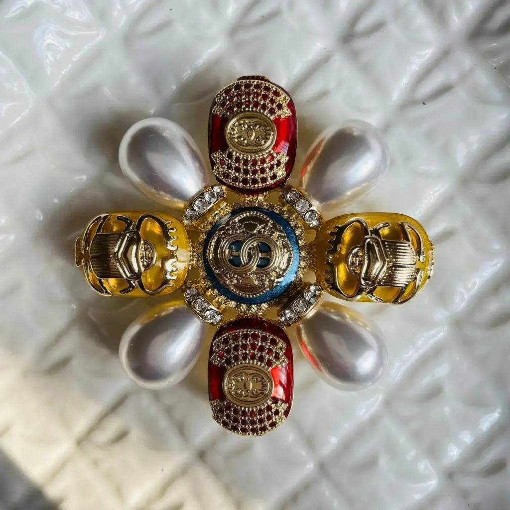 Épingle sertie de rhinestones en forme de coléoptère pour broche de perles designer de luxe Chanel, présentant un médaillon central en émail bleu, avec des éléments entrelacés en or formant le motif CS, entouré de motifs de coléoptères en émail rouge et jaune rehaussés de cristaux et de perles.